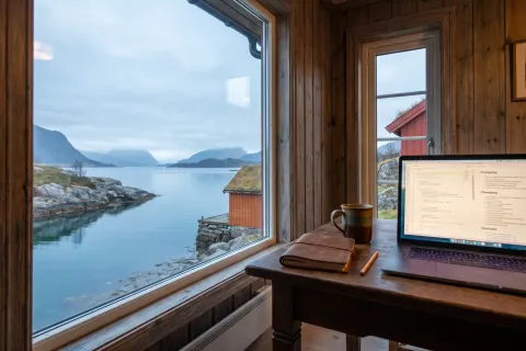 En bærbar datamaskin med en endringslogg på skjermen ligger på et trebord inne i en norsk hytte, med et stort vindu mot en rolig fjord, granittholmer og et okerfarget naust i mykt morgenlys.