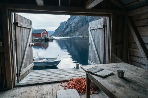 Innsiden av et norsk naust med åpne dører mot en rolig fjord, en liten aluminiumsbåt ved brygga, et rødmalt naust på andre siden av vannet, en åpen feltbok og kaffekopp på et slitt trebord i forgrunnen, og et kveil oransje tau på gulvet
