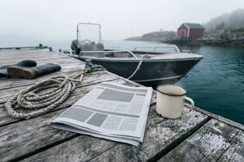 Oppslått avis på en trebrygge ved en norsk fjord, med en kaffekopp ved siden av og en fiskebåt i bakgrunnen i morgenlys